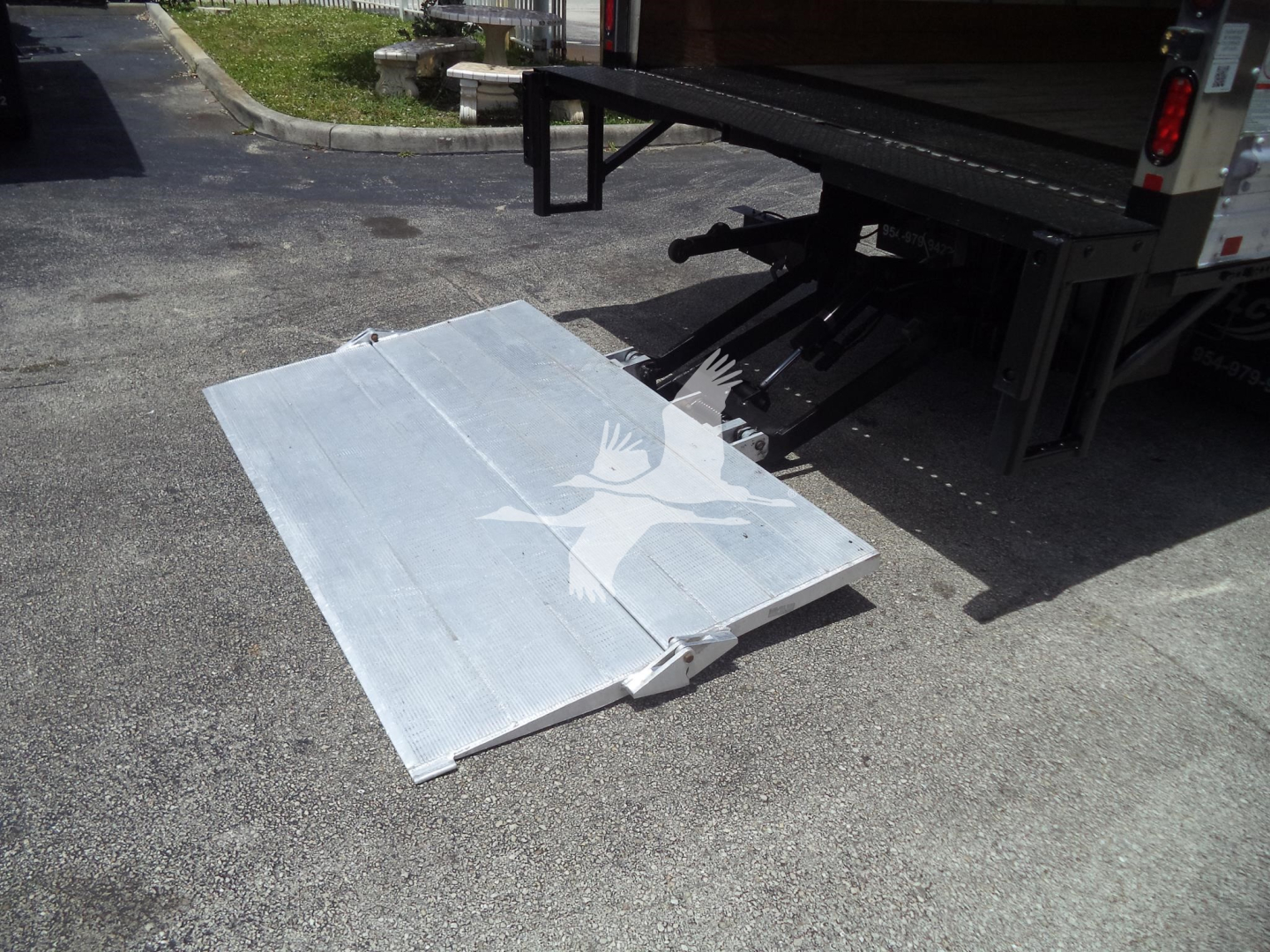 Isuzu NPR HD truck with lowered aluminum loading ramp on pavement; rear of box truck visible.