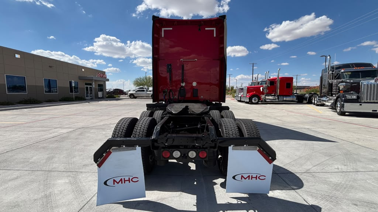 Rear view of a Kenworth truck chassis with red sleeper cab and MHC mud flaps, showing dual rear wheels and hitch area.