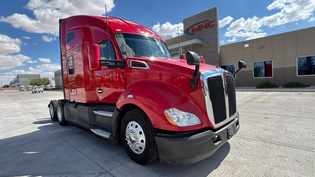 Kenworth T680 truck in red exterior, front-left view, with chrome grille and headlights; dealership building in background; clear blue sky.