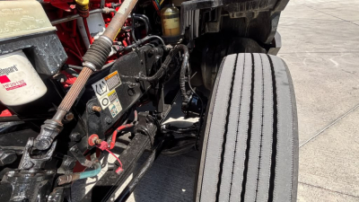 Kenworth truck engine bay area with hoses, wiring, and components visible; plastic reservoirs with labels; front wheel and frame; labels with warning text are partially visible.
