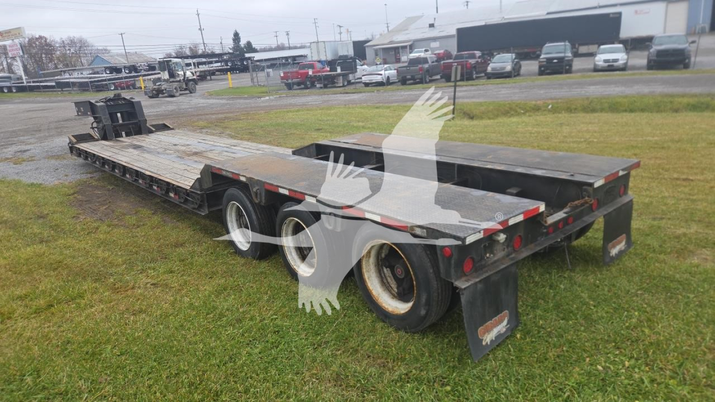Lowboy trailer showing dual rear axles and flatbed deck with metal ramps and wheel wells, used condition.