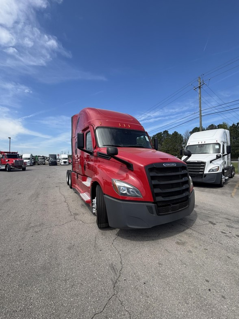 Front-left view of a red Freightliner Cascadia 126 semi-truck in a lot. Visible are the cab with black grille, large bumper, and side mirror. Other trucks in background.