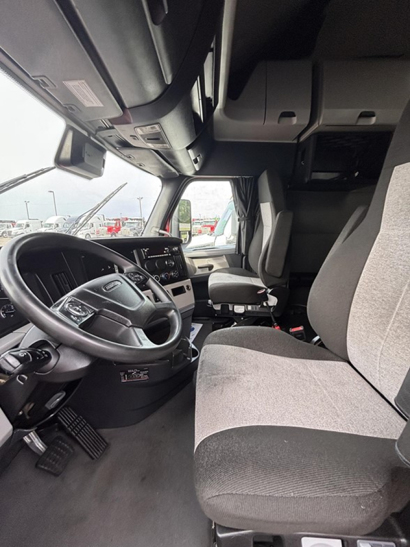 Freightliner Cascadia truck cab interior showing driver's seat, steering wheel, dash, and passenger seat with grey fabric upholstery.