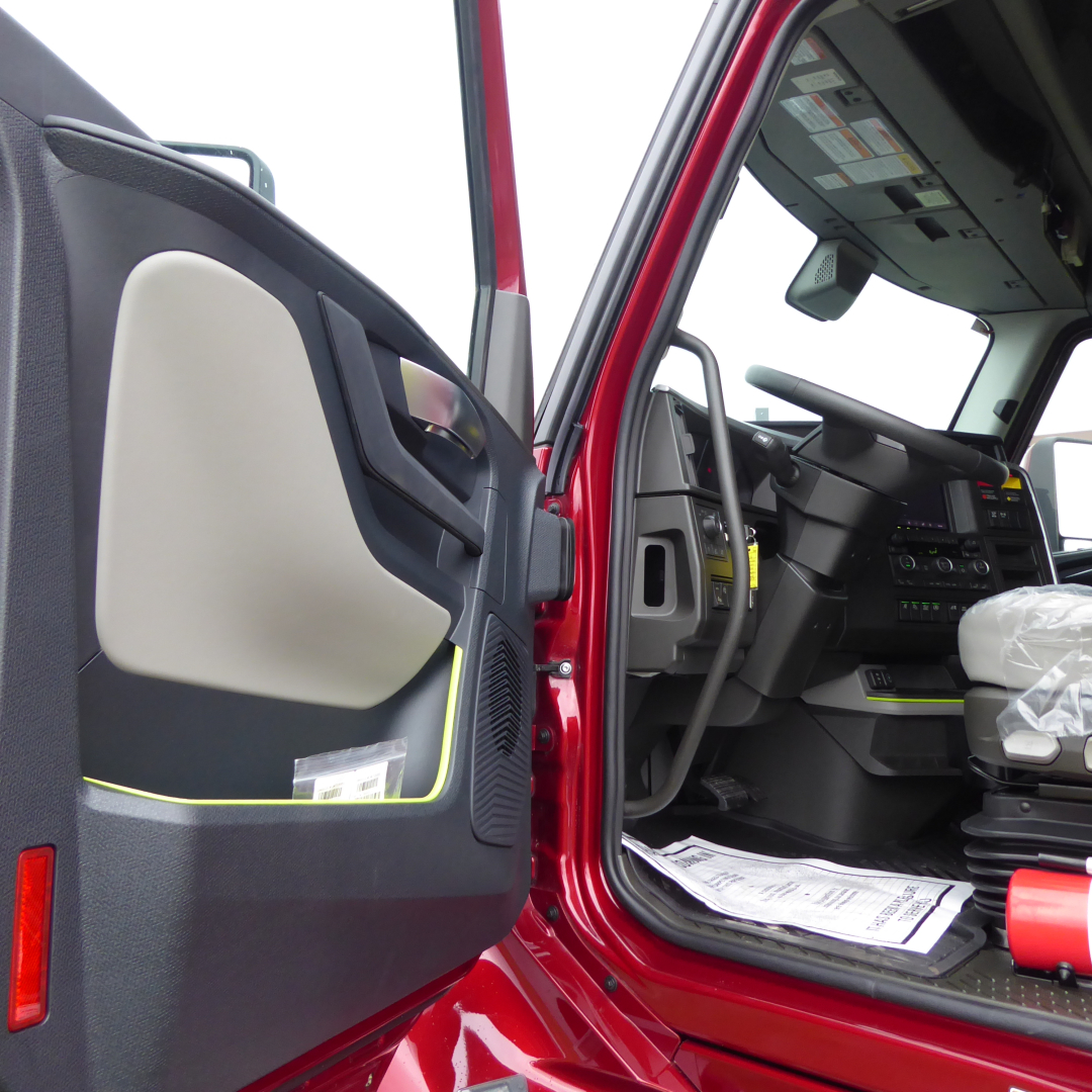 Truck cab interior showing open door and steering column, with dashboard controls and paperwork on the floor; red exterior paint visible.