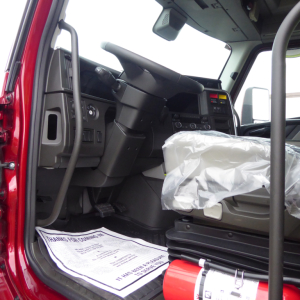 Cab interior of a new Volvo VNL64T860 truck showing the driver seat with plastic cover, dashboard, steering column, pedals, and door frame.