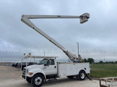 Ford F-750 bucket truck with hydraulic boom raised to extend above cab; white utility body, side storage compartments, bucket at end of arm.