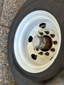 Wheel and hub assembly of a trailer with white steel rim, tire, and central hub with cables visible.