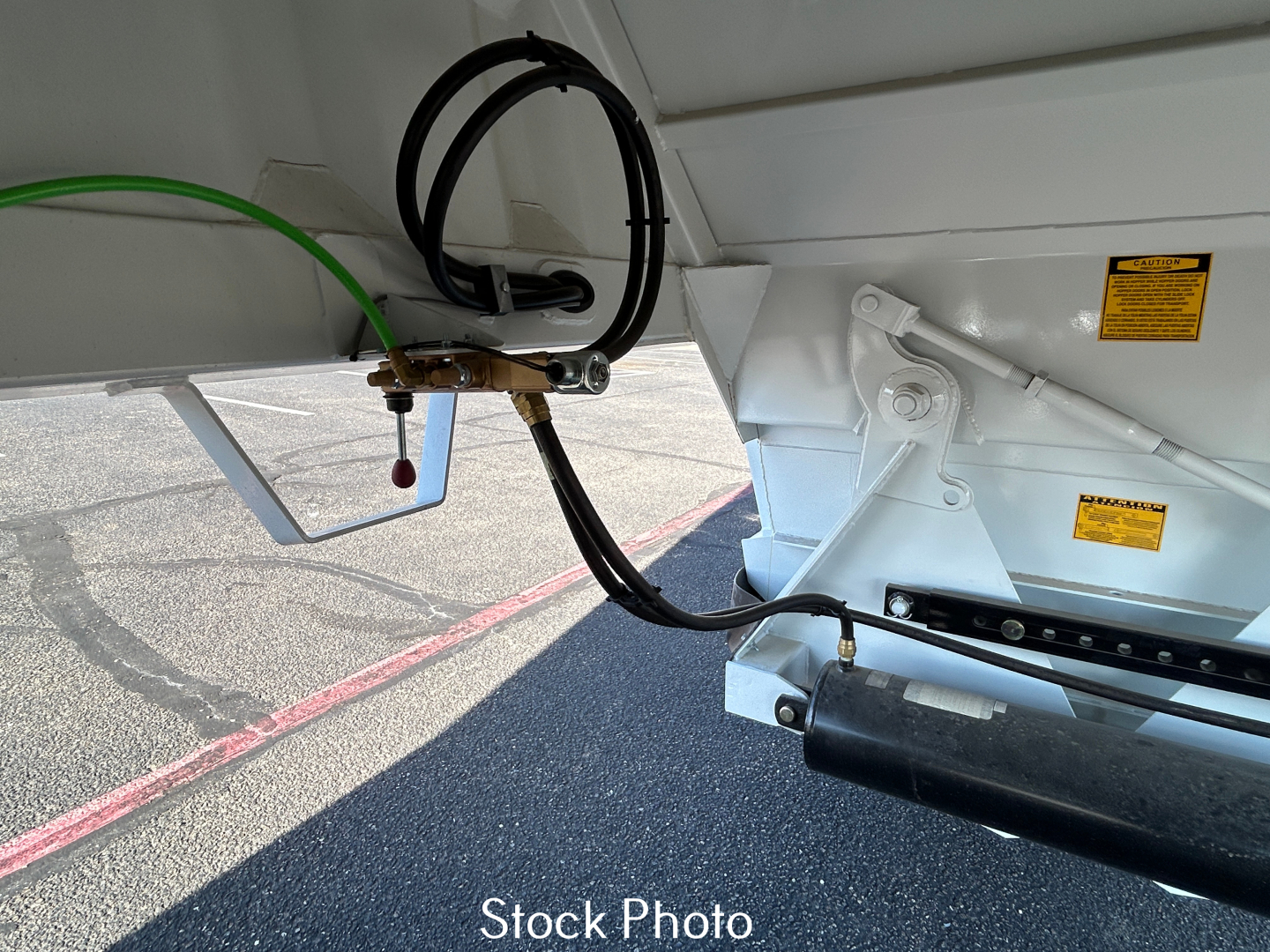 Trailer hitch area showing hydraulic lines and frame with stickers, black roller bar, and green hose connected to a spring-dampened mechanism.