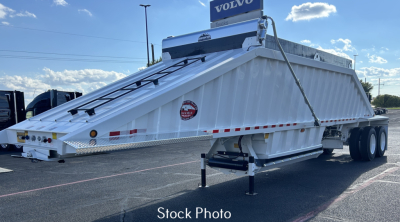 Trailer dump bed in parking lot with Volvo sign in background, side view showing white steel dump trailer with hydraulic mechanism and safety stickers.