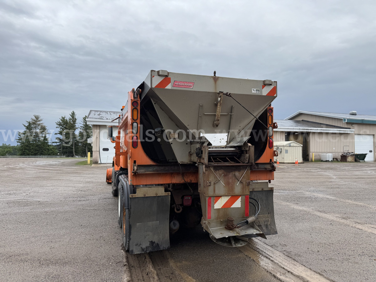 Dump truck rear with spread hopper, tailgate and warning chevrons visible. Industrial facility in background.