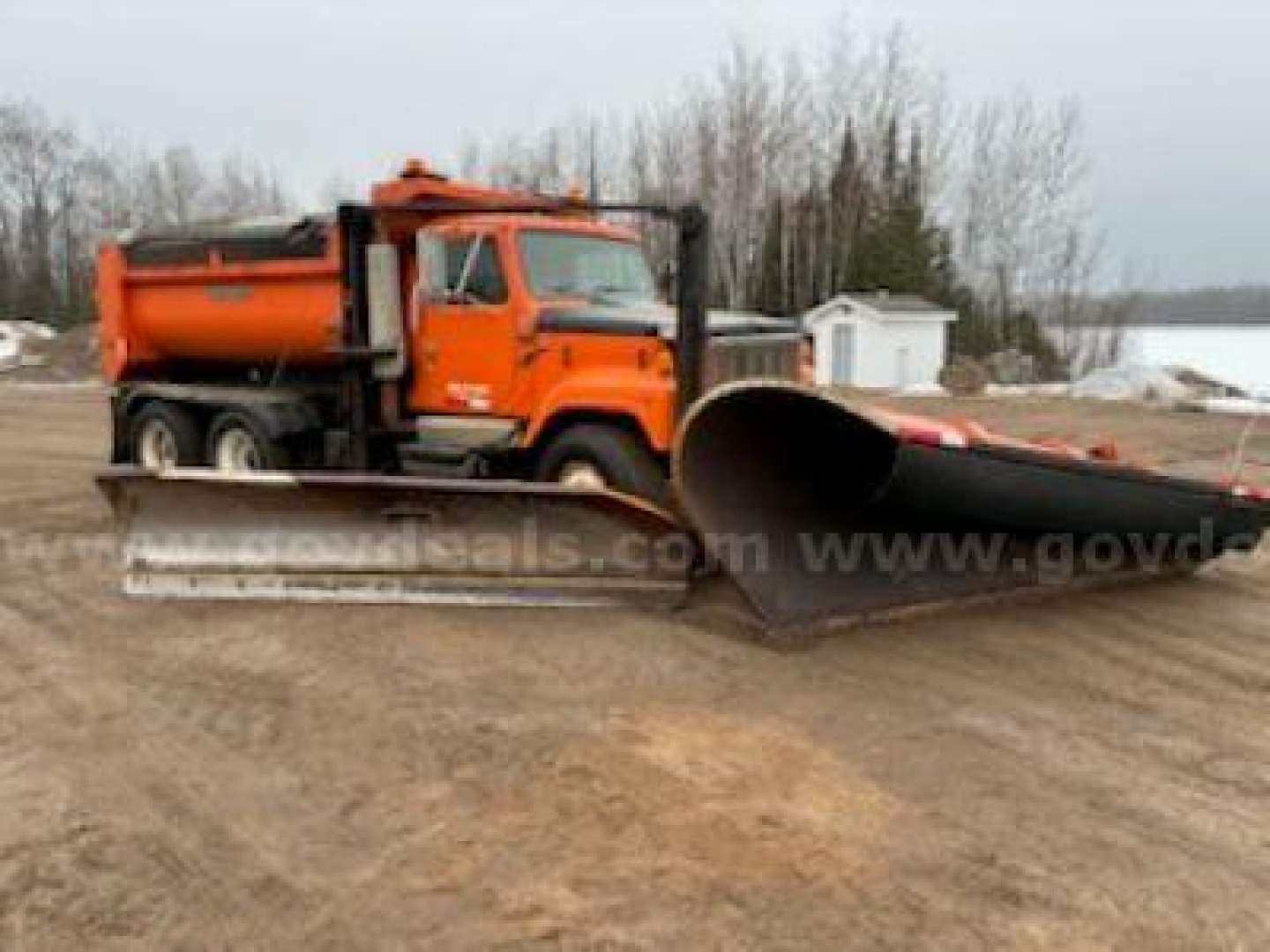 Dump truck with large orange snowplow blade attachment in foreground.