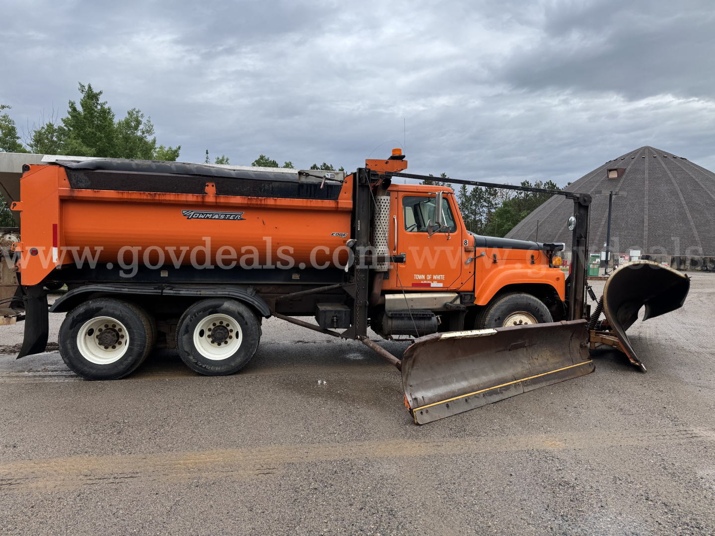 Orange International dump truck with snow plow blade in front, large black-topped bed, two rear axles, and white wheels. Side text shows truck model details and stock number; the image may include a watermark from govdeals.