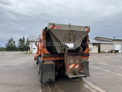 Dump truck rear with spread hopper, tailgate and warning chevrons visible. Industrial facility in background.