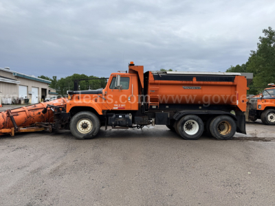 Dump truck side view showing orange International dump truck with black dump bed and snow plow attachment. Visible text on door includes 'INTERNATIONAL' and '2574' with stock/ID stickers.