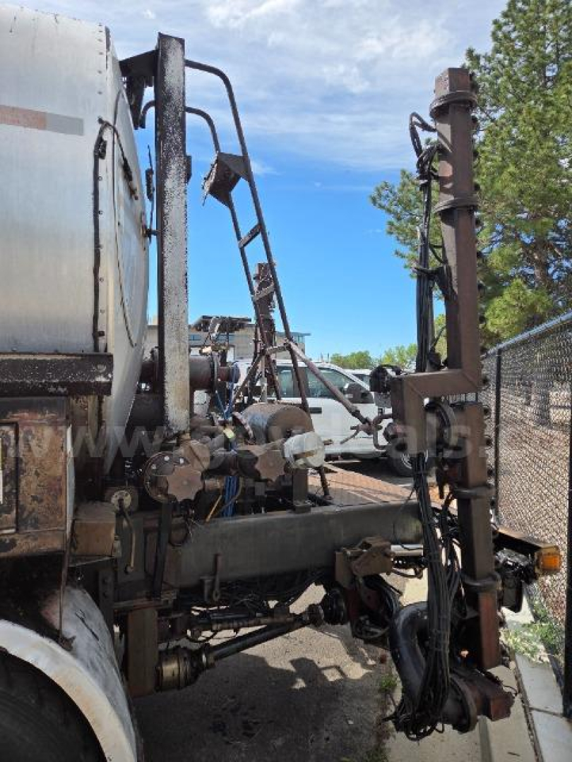Truck equipment with hydraulic hoses and a vertical lifting apparatus attached to a trailer or truck chassis. Parts show metal framework, hoses, and a rusty vertical post.