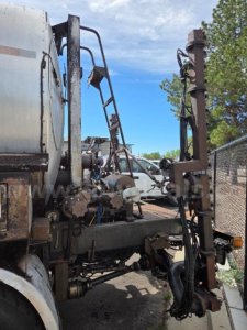 Truck equipment with hydraulic hoses and a vertical lifting apparatus attached to a trailer or truck chassis. Parts show metal framework, hoses, and a rusty vertical post.