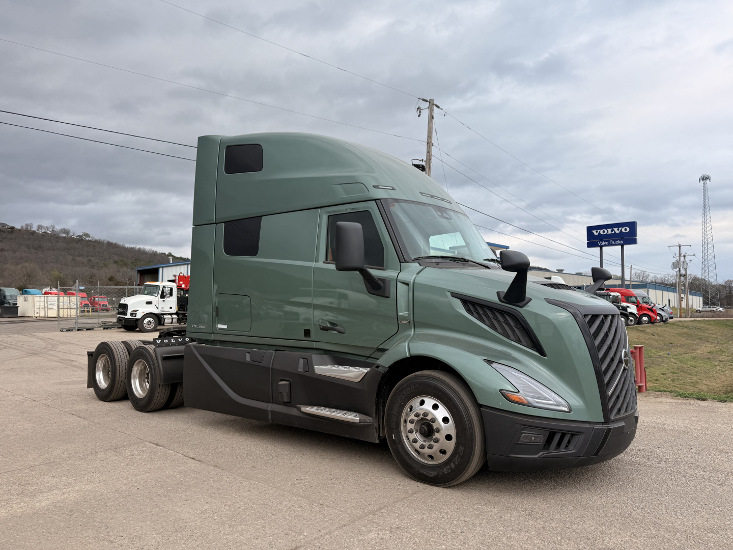 Volvo VNL64T860 semi-truck tractor in a dealership lot, green cab with sleeper, three rear axles, Volvo sign in background.