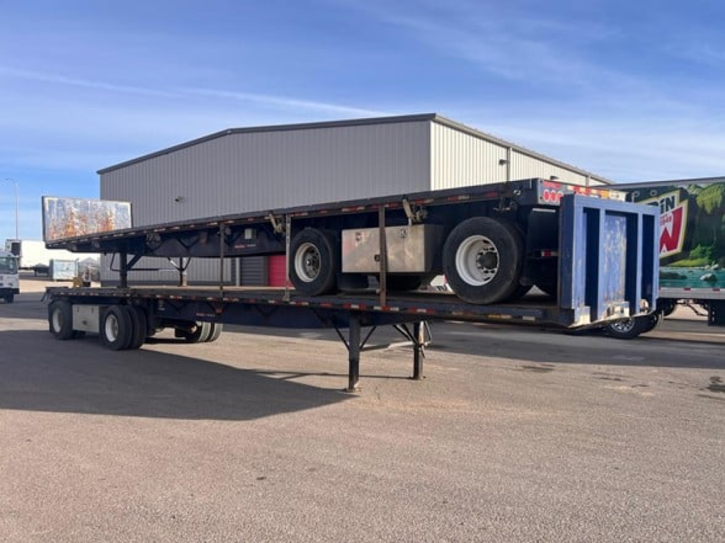 Flatbed trailer with a Great Dane Freedom Se model on a lowboy, showing dual rear axles, spare tire, and a blue ramp at the back.