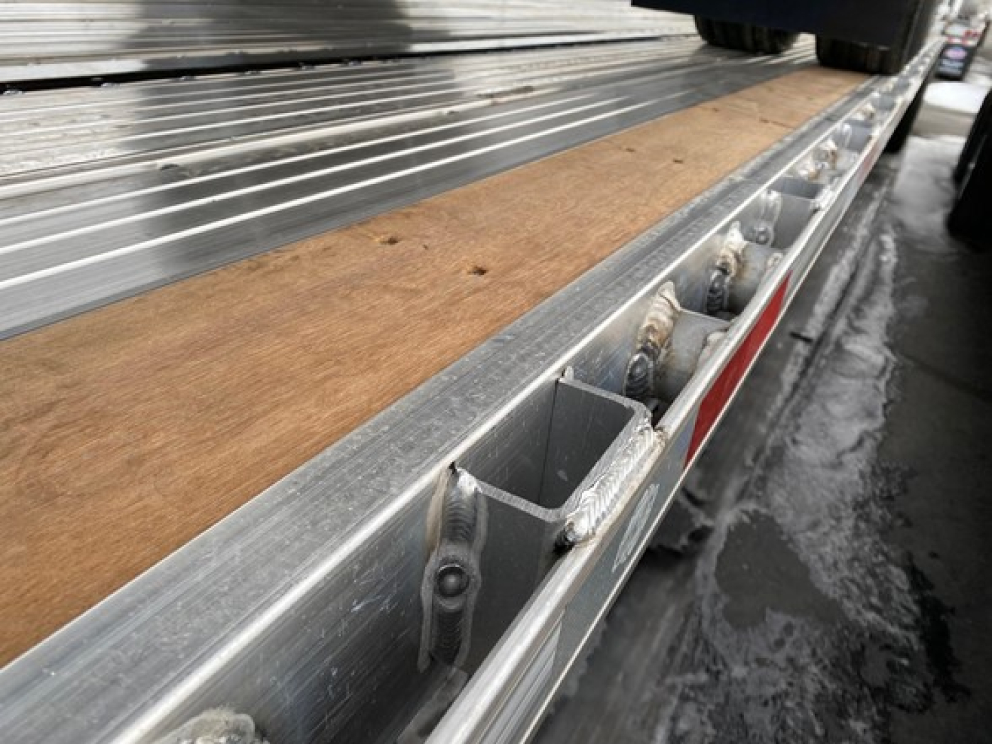 Flatbed trailer side rail with wooden deck and aluminum edge rails, showing stake pockets and metal flooring