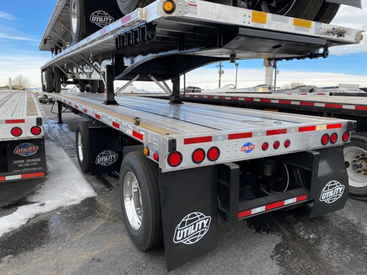 Utility trailer with flatbed sections and a lift deck, showing aluminum deck, rear lights, mud flaps with Utility logo, dual rear axles, and a spare tire mounted under deck.