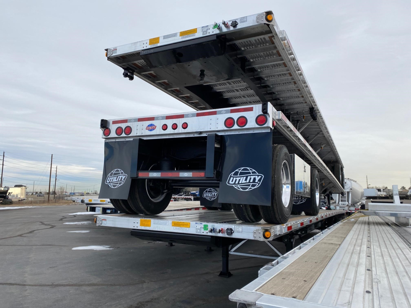 Utility 4000AE flatbed trailer on a loading dock with the bed raised, dual rear axles with black wheels and white rims, reflective lights, and logo decals, background with overcast sky.