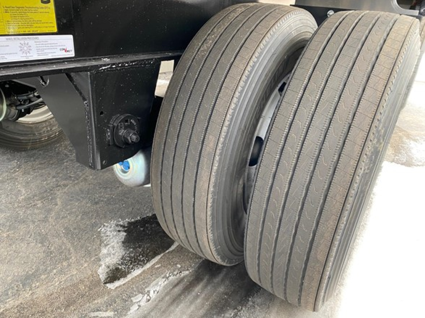 Trailer rear axle with dual tires and black frame; shows side view of two straight-axle tires mounted under a flatbed trailer, wheel hub and mounting bolts visible.