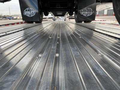 Trailer bed of a 2024 Utility 4000AE flatbed trailer showing corrugated metal deck with tie-down anchors and rear wheels visible in background.