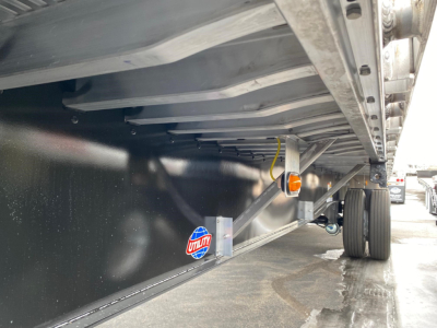 Underbelly of a new Utility 4000AE flatbed trailer showing frame, side rail, and spare hubs; reflective sticker visible along the black underside.