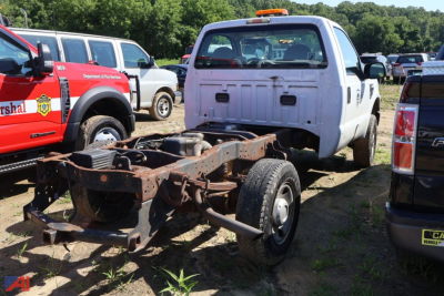 Truck chassis and cab frame in a salvage yard with white cab and exposed wheelbase; other trucks parked nearby