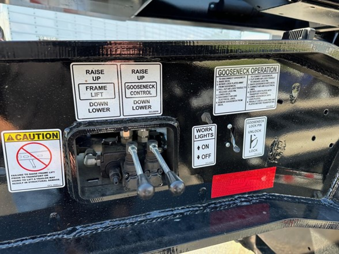 Trailer control panel with labels: 'RAISE UP FRAME LIFT DOWN LOWER', 'GOOSENECK CONTROL DOWN UP', 'WORK LIGHTS ON OFF', and caution sign; levers and warning decals on black metal surface.
