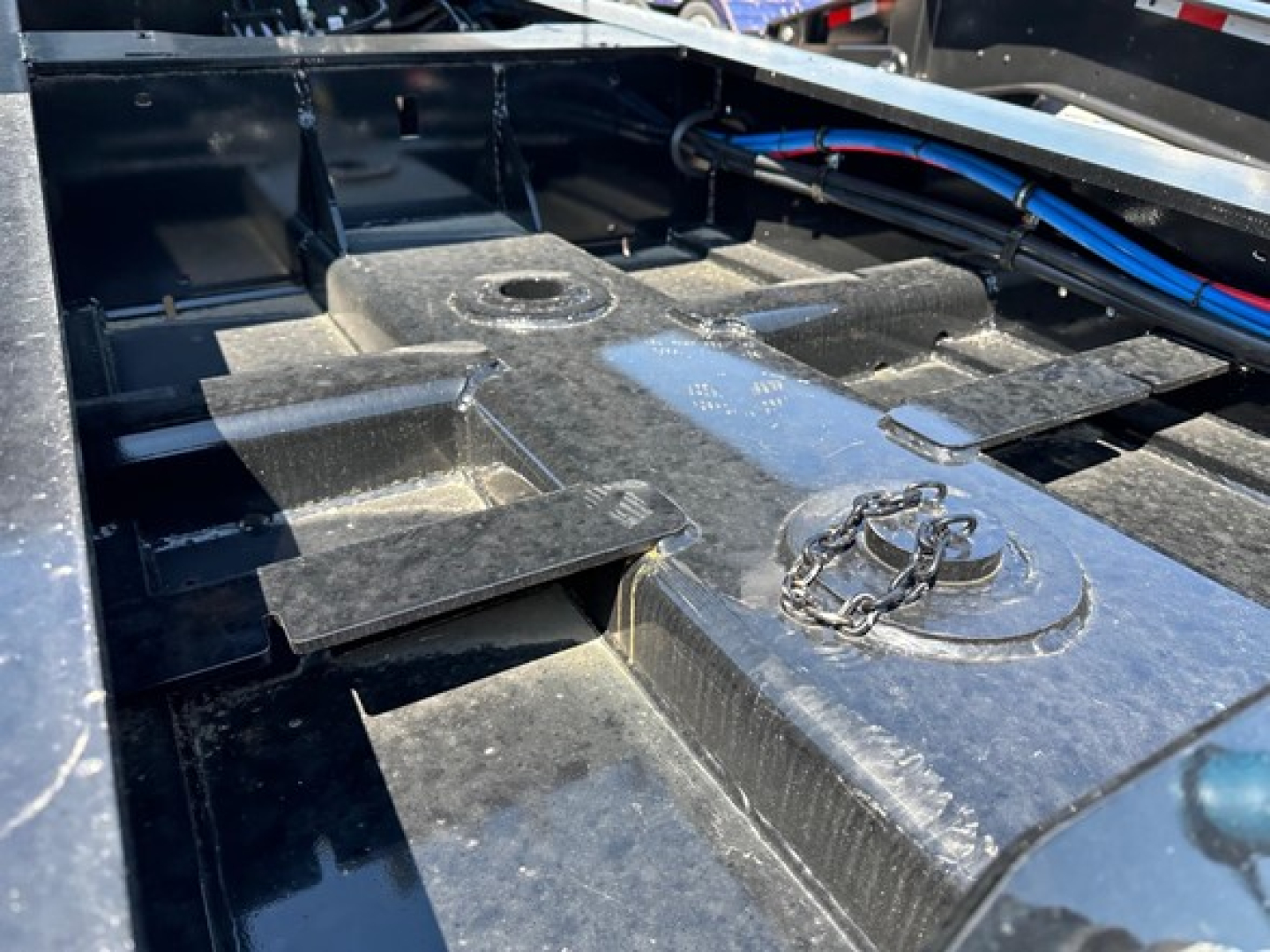Trailer deck with metal frame and hydraulic lines; visible components include a central mounting pad with a chained ring and a circular hole, plus blue and black hoses along the top edge.