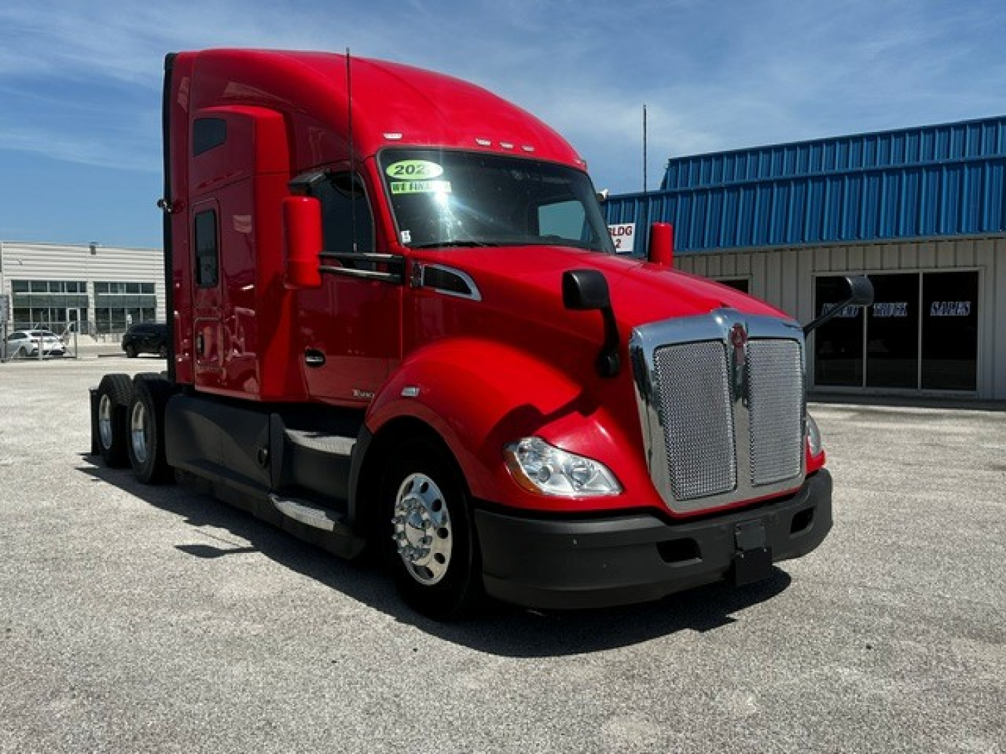 Kenworth T680 tractor-trailer in red, front-left view, with chrome grille, daytime running lights, and blue industrial building in background.