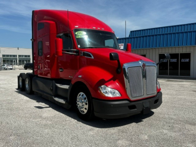 Kenworth T680 tractor-trailer in red, front-left view, with chrome grille, daytime running lights, and blue industrial building in background.