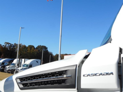 Front portion of a white Freightliner Cascadia 126 cab showing Cascadia badge and grille, view of side with door frame and hood.