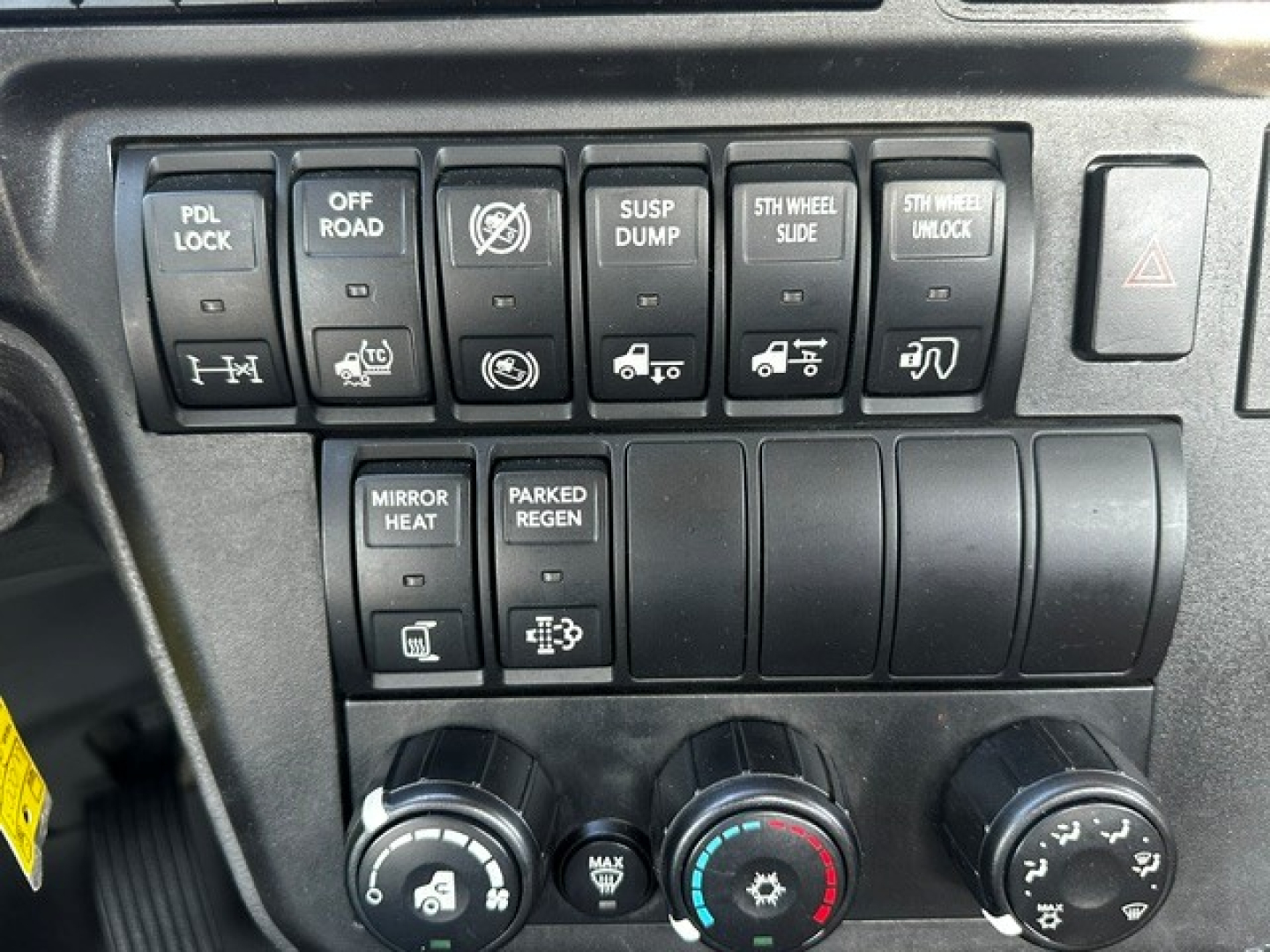 Dashboard control panel with multiple switches and climate knobs in a truck cab. Visible labels include PDL LOCK, OFF ROAD, SUSP DUMP, 5TH WHEEL SLIDE, 5TH WHEEL UNLOCK, MIRROR HEAT, PARKED REGEN, and three climate control dials.