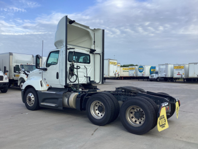 Truck side view showing 2021 International LT625 conventional daycab with extended sleeper hood raised, dual rear axles, and fifth wheel, in a lot with other trucks visible.