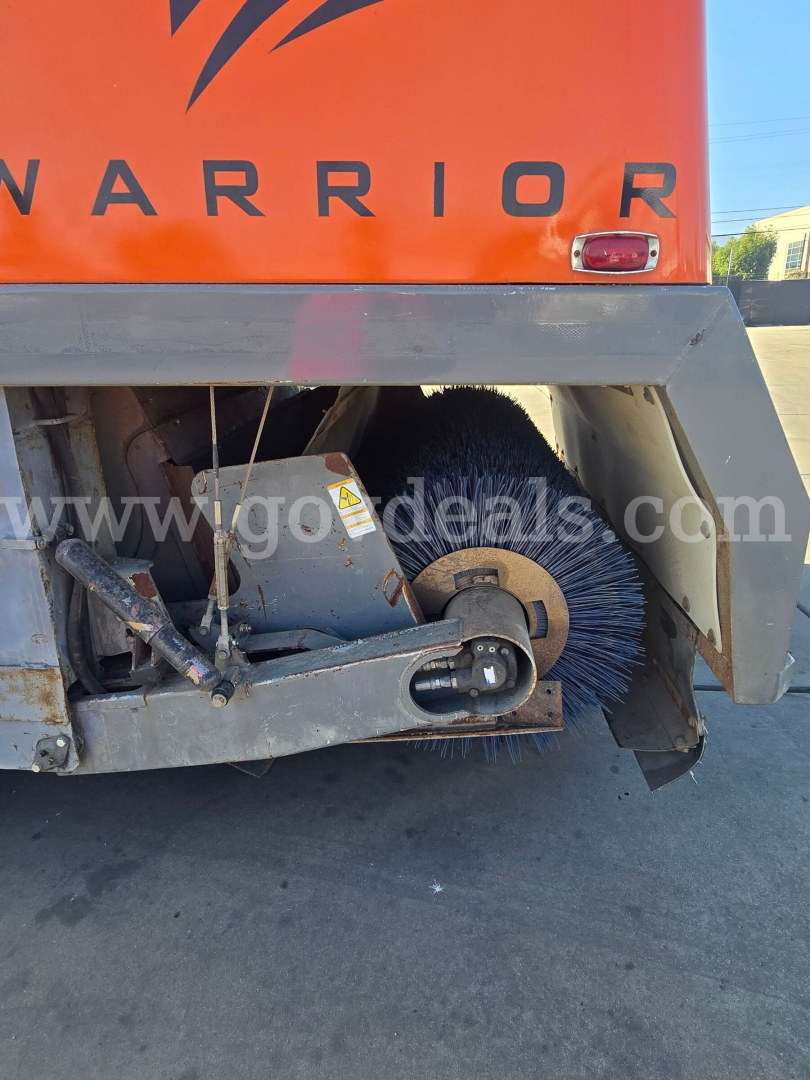 Freightliner M2 112 street sweeper rear showing side turning brush and vacuum housing with Warrior branding; hydraulic linkage and protective framework visible.