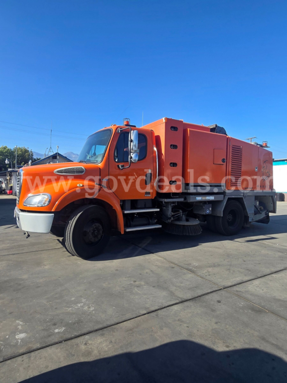 Orange Freightliner M2 112 street sweeper truck in a parking lot, side view showing cab, sweeping brushes under the front, and large equipment body in back with vents and panels.