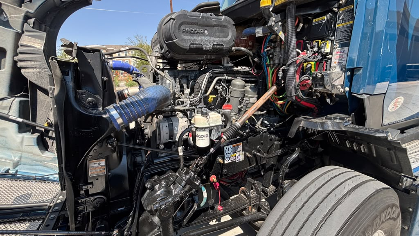 Kenworth T680 truck engine bay showing exposed components, hoses, and wiring harnesses; air intake and various fluid reservoirs visible.