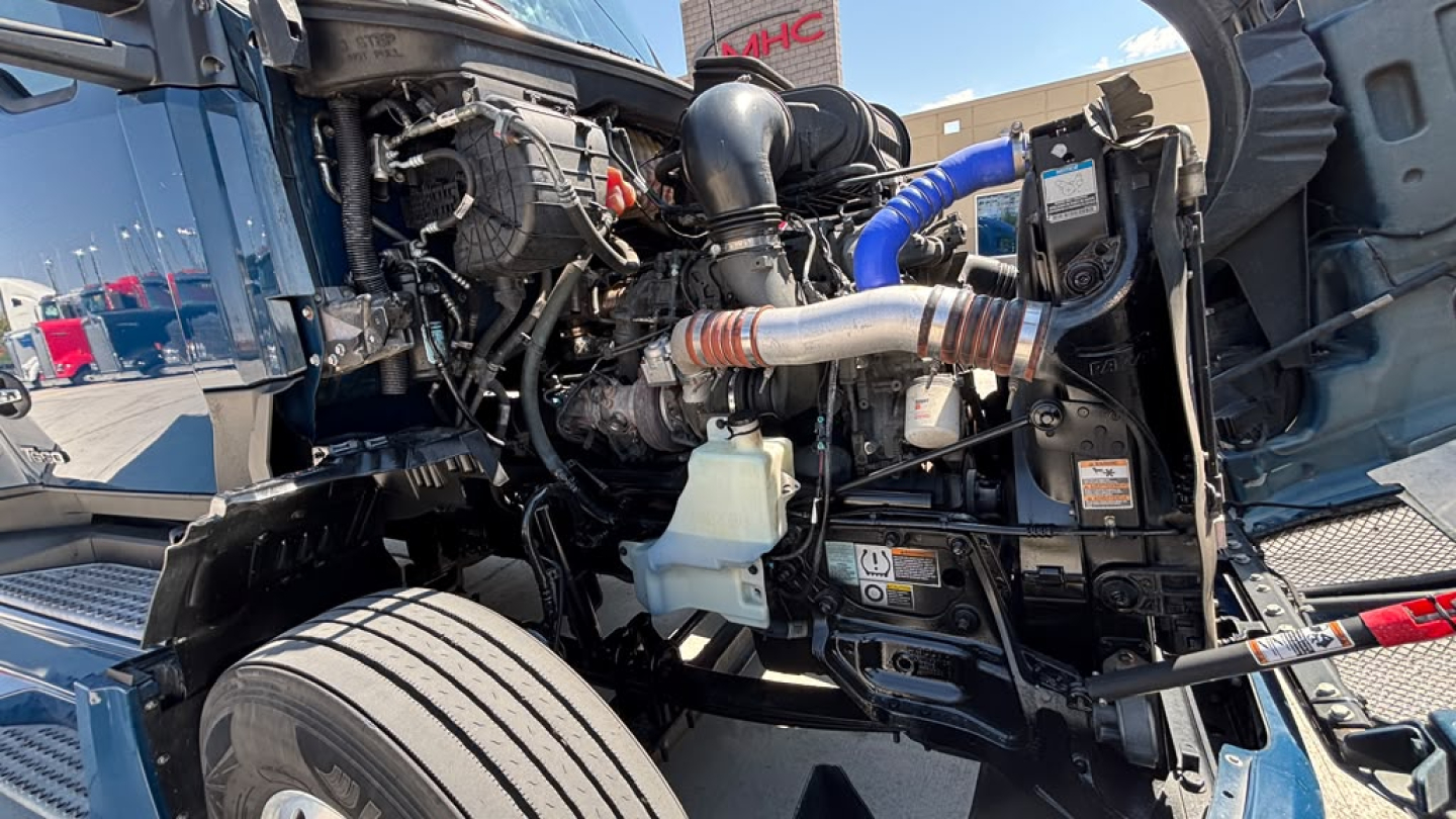 Kenworth T680 engine bay showing hoses, clamps, coolant reservoir, and components in a truck with exposed hood, front wheel, and radiator area.