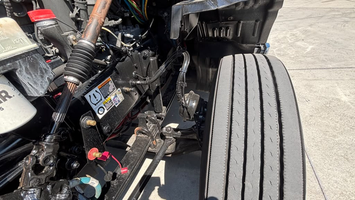 Kenworth T680 engine bay with visible fluid reservoirs and wiring; front right wheel visible; labels and warning stickers on nearby components.