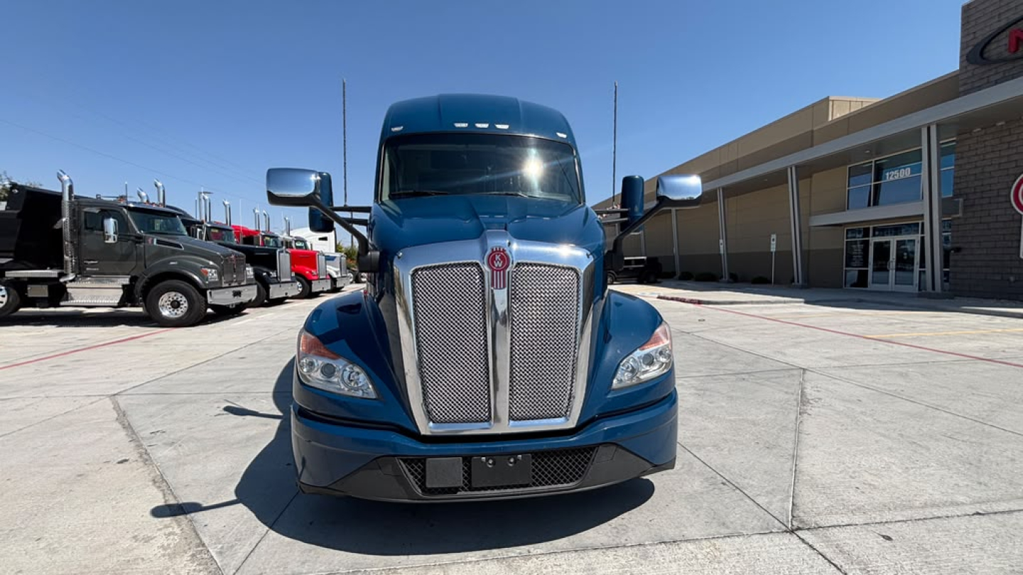 Kenworth T680 front view showing large chrome grille and headlights, blue cab, side mirrors, and parking lot backdrop.
