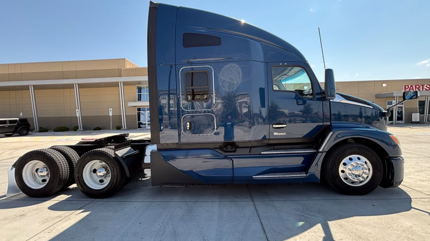 Truck side view of a blue 2023 Kenworth T680 conventional sleeper with dual rear axles and chrome wheels.