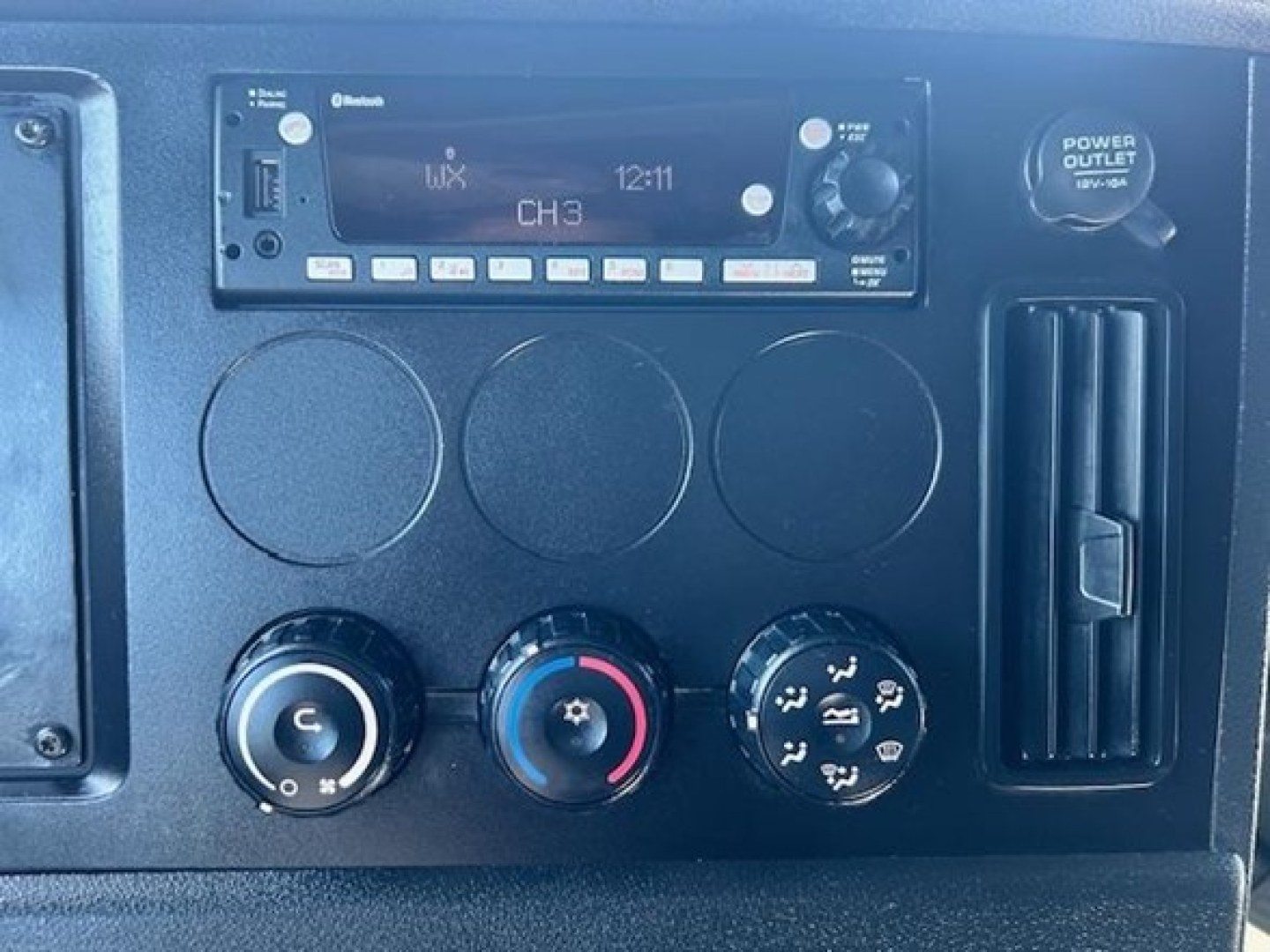 Dashboard panel of a Freightliner Cascadia 126 showing infotainment unit with CH3, three circular knobs for climate control, and a power outlet/12V socket.