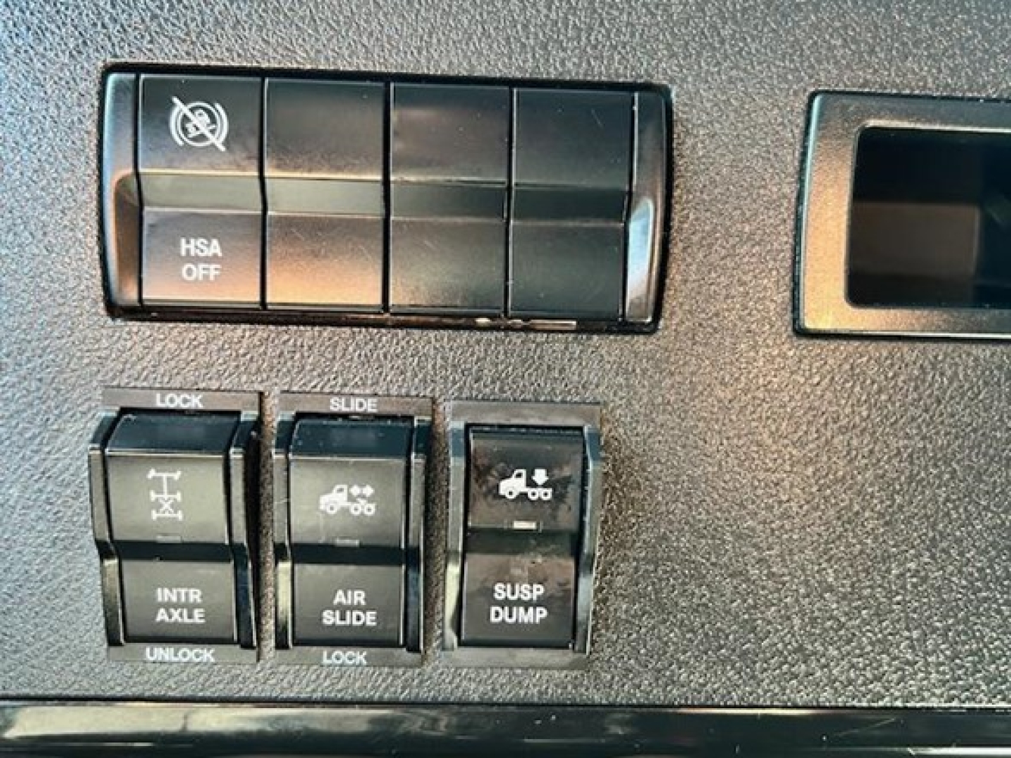 Dashboard switches and labels in a Freightliner Cascadia cab: HSA OFF switch area, and row of levers labeled LOCK, SLIDE, INTR AXLE, AIR SLIDE, SUSP DUMP.