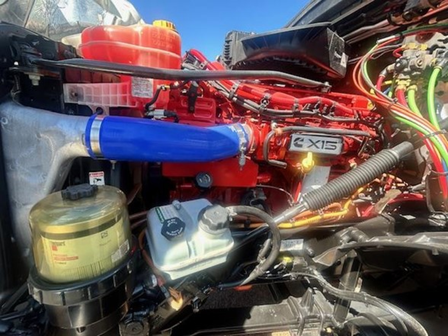 Freightliner Cascadia engine bay with bright red engine, blue intercooler hose, various fluid reservoirs, and wiring harnesses visible.