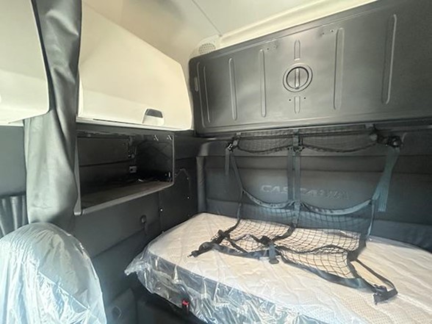 Freightliner Cascadia sleeper cab interior showing bed with mesh storage, side shelves, and overhead cabinet; window shades and interior panels visible.