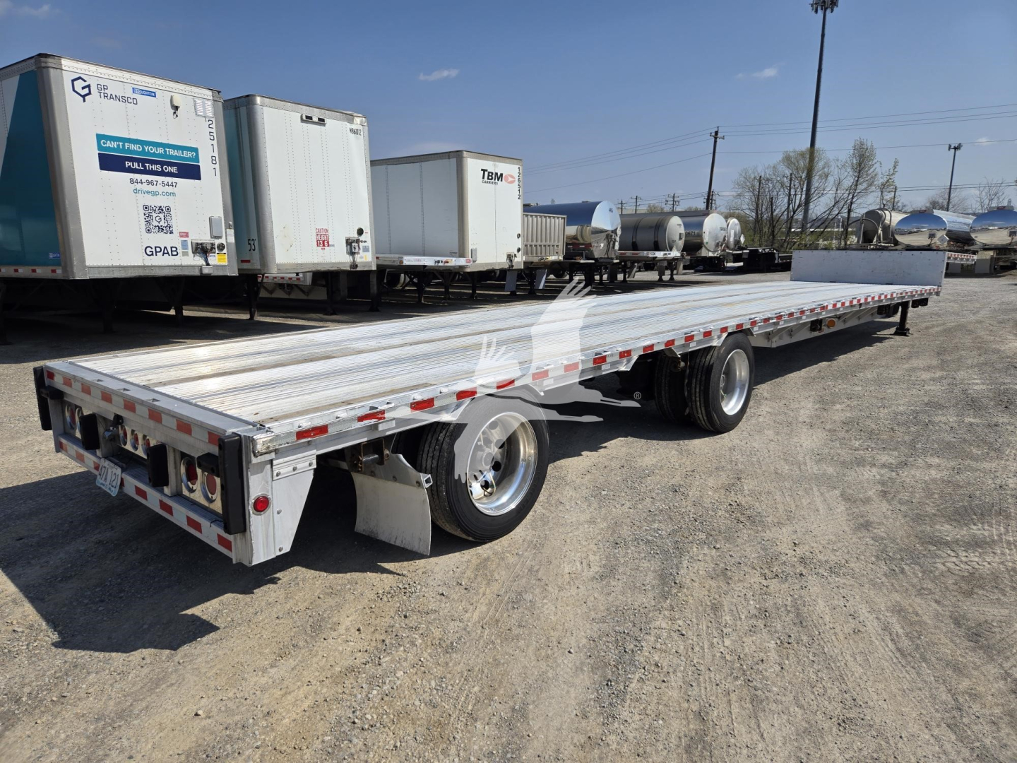 Flatbed trailer in a used equipment lot showing long aluminum deck with dual rear axle and wheel fenders; drop deck trailer with red-and-white reflective tape