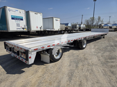Flatbed trailer in a used equipment lot showing long aluminum deck with dual rear axle and wheel fenders; drop deck trailer with red-and-white reflective tape