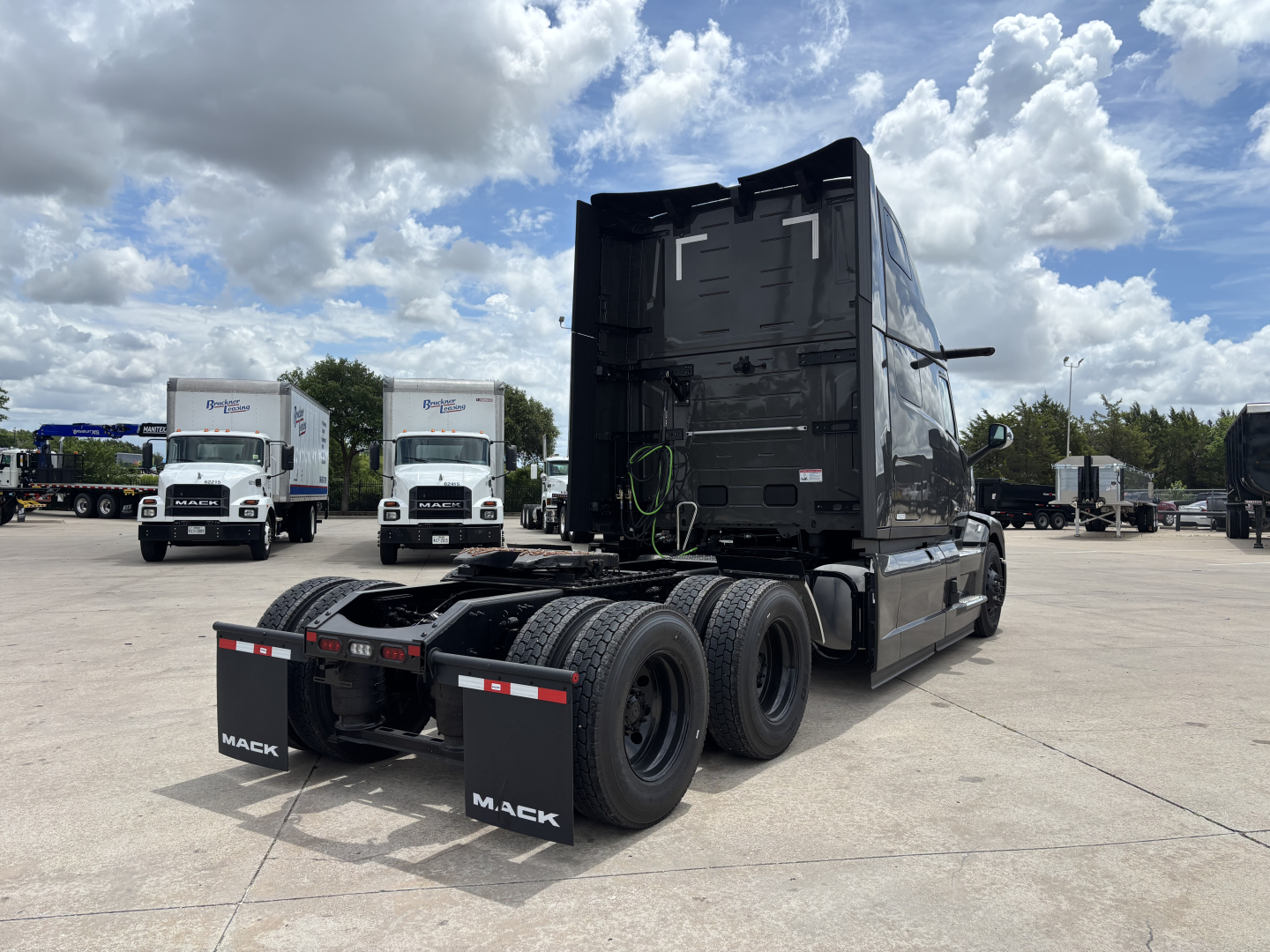 Rear three-quarter view of a new 2027 Mack PR64T76H conventional sleeper truck. The tractor has a high-roof sleeper, tandem rear axles, black paint, and mud flaps with Mack branding; visible fuel/wheel areas and fifth wheel; clear sky backg